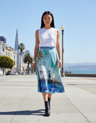 Woman wearing a white top and blue patterned skirt standing on a city street with a scenic background.