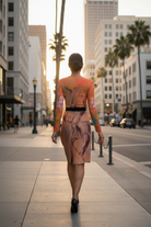 Woman in a colorful dress walking on a city street with tall buildings and palm trees in the background.