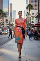 Woman in a colorful dress walking on a city street with tall buildings and palm trees in the background.