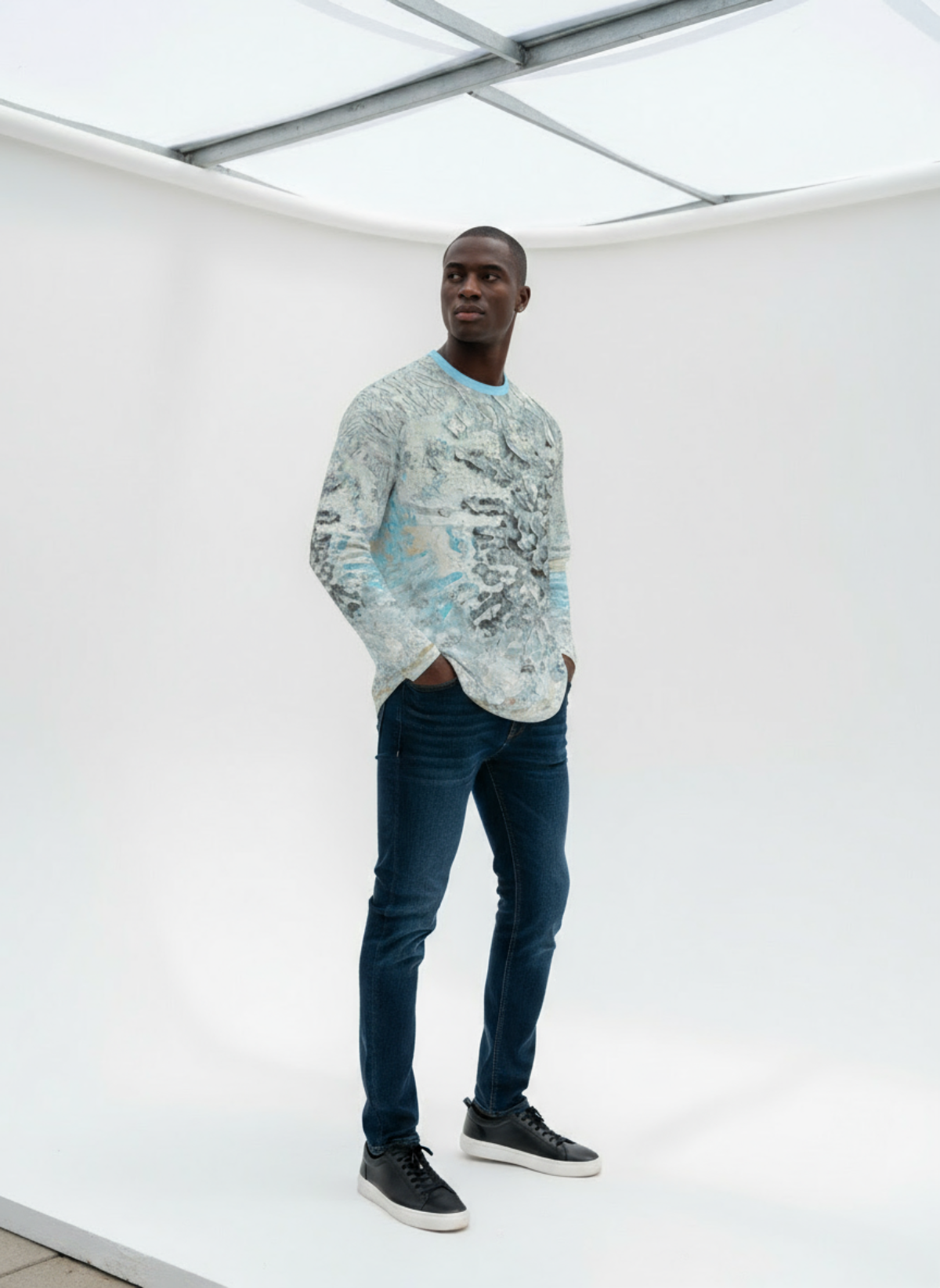 Man wearing a patterned sweater and jeans standing in a minimalistic indoor setting.