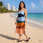Woman in a colorful striped dress standing on a beach with palm trees and buildings in the background.