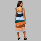 Woman in a colorful striped dress standing on a sunlit street with pastel-colored buildings.