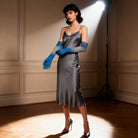 Woman in a gray satin dress and opera gblue gloves standing in a spotlight against a beige wall.